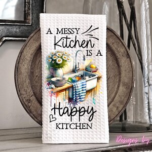 May include: White waffle-weave kitchen towel with the text "A Messy Kitchen is a Happy Kitchen." The towel features a watercolor illustration of a kitchen sink with dishes, flowers, and a striped towel. The design includes a heart symbol.