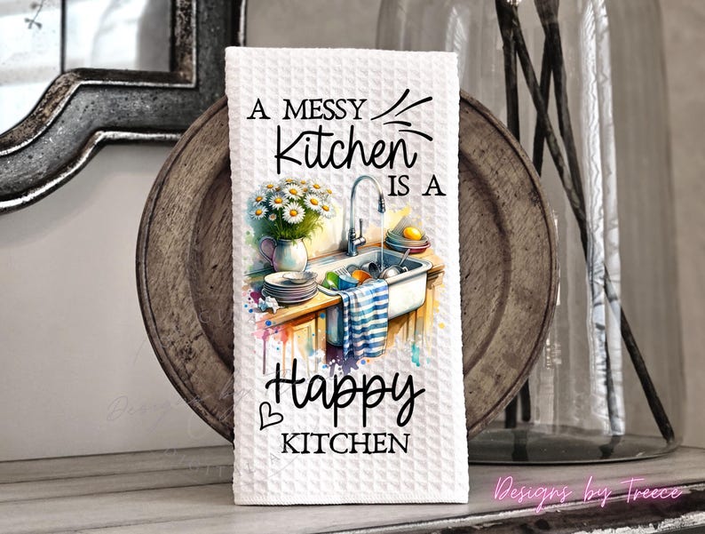 May include: White waffle-weave kitchen towel with the text "A Messy Kitchen is a Happy Kitchen." The towel features a watercolor illustration of a kitchen sink with dishes, flowers, and a striped towel. The design includes a heart symbol.