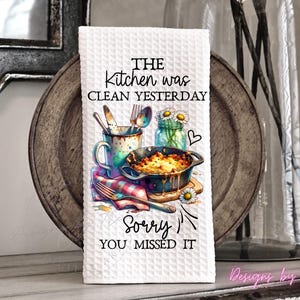 May include: White kitchen towel with a waffle weave texture. The towel features a colorful illustration of kitchen utensils, a casserole dish, and a jar of daisies. Text on the towel reads "THE kitchen was clean yesterday, Sorry YOU MISSED IT."