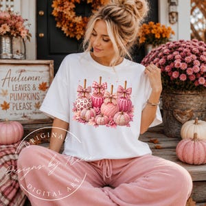 May include: A white short-sleeve t-shirt featuring a graphic print of several pink pumpkins and candy apples decorated with bows, leopard print, and plaid patterns, accented with small pink leaves and sparkles on the front center.