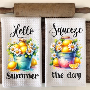 Puede incluir: Dos paños de cocina blancos con tejido de gofre con ilustraciones de acuarela de limones en cubos con margaritas. Una toalla dice "Hello Summer" y la otra "Squeeze the day". Un rodillo de madera y una tabla de cortar están en el fondo.