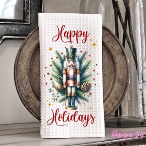 May include: A white waffle-textured kitchen towel featuring a central graphic of a nutcracker surrounded by pine branches, pinecones, and red berries. The text Happy Holidays is printed in red script above and below the nutcracker illustration.