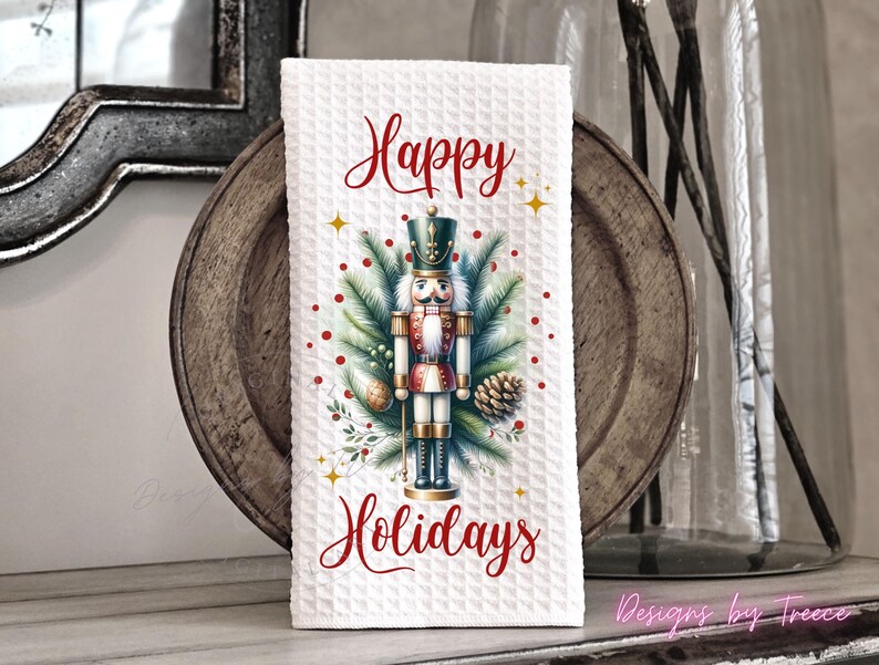 May include: A white waffle-textured kitchen towel featuring a central graphic of a nutcracker surrounded by pine branches, pinecones, and red berries. The text Happy Holidays is printed in red script above and below the nutcracker illustration.
