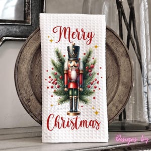 May include: A white waffle-textured kitchen towel featuring a central illustration of a nutcracker soldier surrounded by green pine branches and red berries. The text Merry Christmas is printed in red script above and below the nutcracker figure.