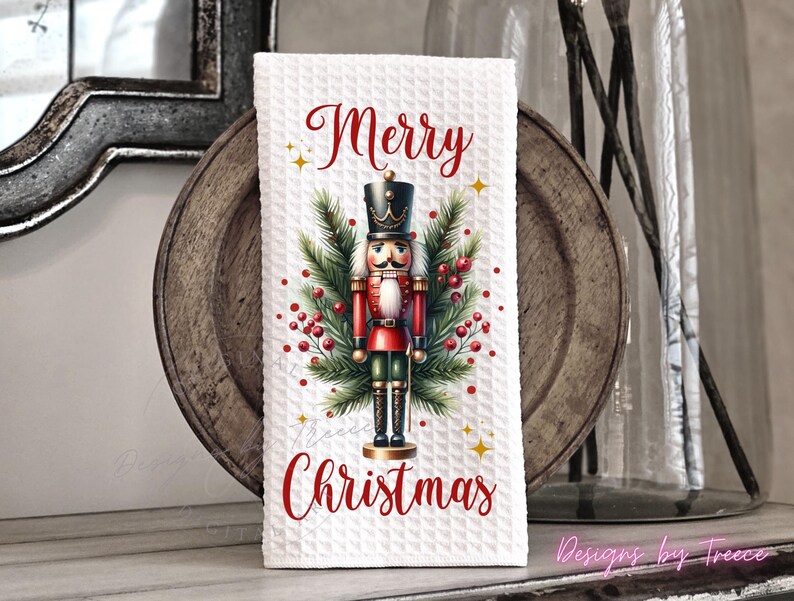May include: A white waffle-textured kitchen towel featuring a central illustration of a nutcracker soldier surrounded by green pine branches and red berries. The text Merry Christmas is printed in red script above and below the nutcracker figure.