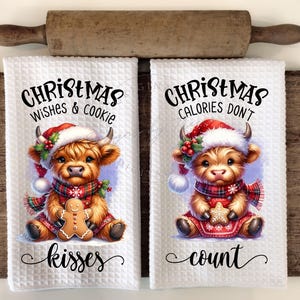 Op de afbeelding: Twee witte keukendoeken met een rood-wit Highland-koe-ontwerp. Een handdoek zegt "Christmas Wishes & Cookie Kisses" en de andere zegt "Christmas Calories Don't Count".