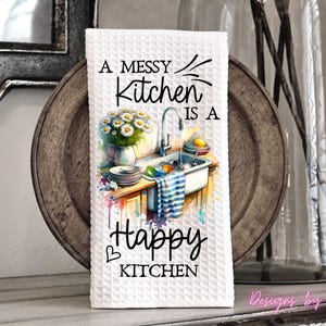 May include: White waffle-weave kitchen towel with the text "A Messy Kitchen is a Happy Kitchen." The towel features a watercolor illustration of a kitchen sink with dishes, flowers, and a striped towel. The design includes a heart symbol.
