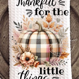 May include: White waffle-weave kitchen towel with a plaid pumpkin design. The pumpkin is surrounded by fall foliage and flowers. Text reads "Thankful for the little things" in black script.