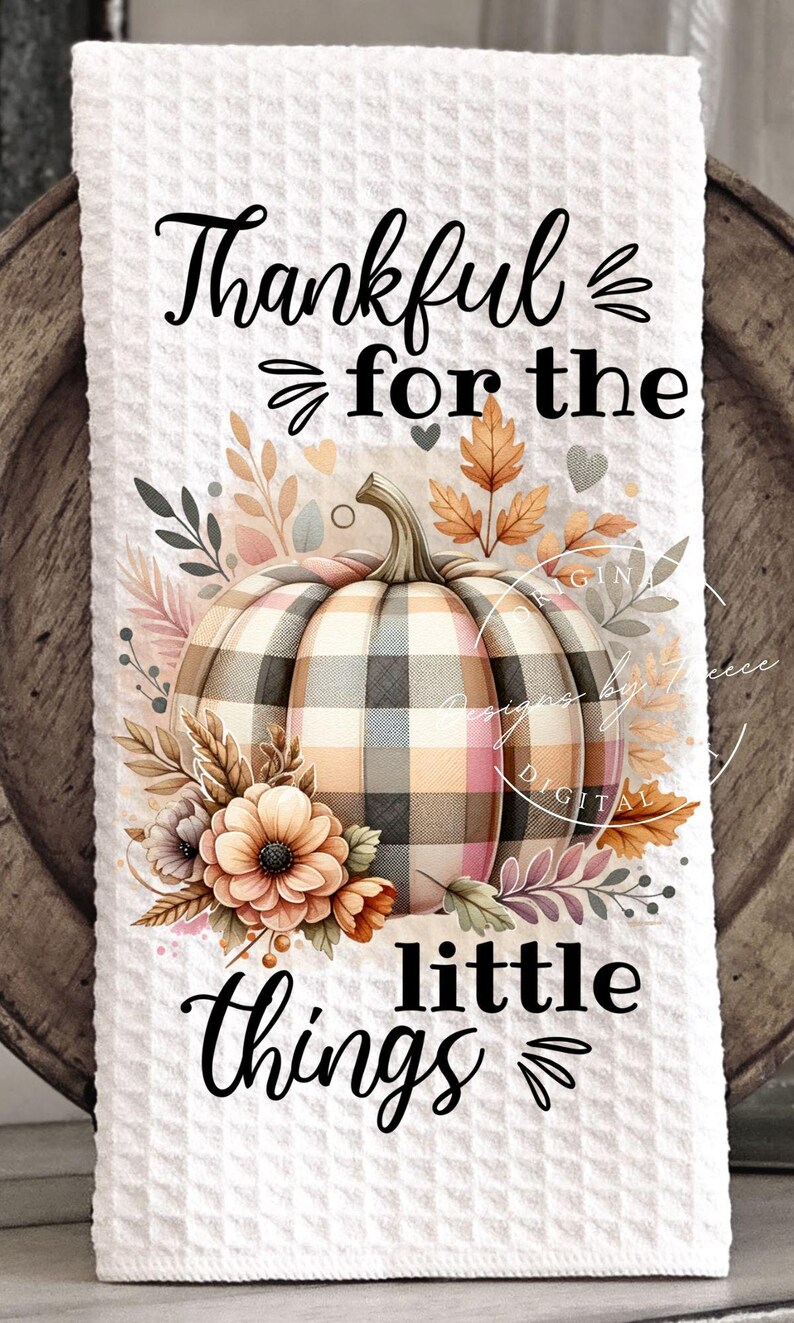 May include: White waffle-weave kitchen towel with a plaid pumpkin design. The pumpkin is surrounded by fall foliage and flowers. Text reads "Thankful for the little things" in black script.
