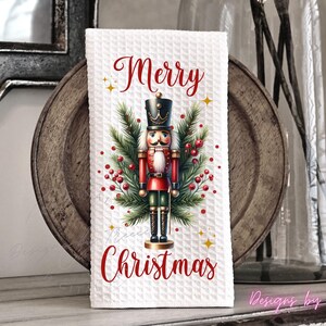 May include: A white waffle-textured kitchen towel featuring a central illustration of a nutcracker soldier surrounded by green pine branches and red berries. The text Merry Christmas is printed in red script above and below the nutcracker figure.