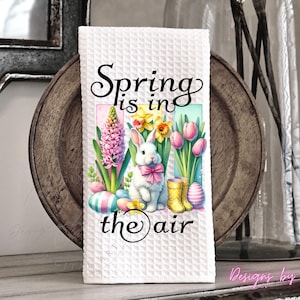 May include: White waffle-weave kitchen towel with a colorful spring-themed design. The design features a white bunny with a pink bow, flowers, Easter eggs, and the text "Spring is in the air."