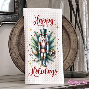 May include: A white waffle-textured kitchen towel featuring a central graphic of a nutcracker surrounded by pine branches, pinecones, and red berries. The text Happy Holidays is printed in red script above and below the nutcracker illustration.