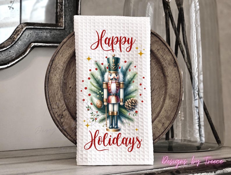 May include: A white waffle-textured kitchen towel featuring a central graphic of a nutcracker surrounded by pine branches, pinecones, and red berries. The text Happy Holidays is printed in red script above and below the nutcracker illustration.