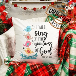 Könnte beinhalten: Weißes quadratisches Kissen mit dem Text "I WILL SING OF THE goodness OF God" und Psalm 89:1. Das Kissen zeigt ein Design von vier bunten Vögeln, die singen und auf einem Nest sitzen. Das Kissen liegt auf einem Stuhl mit einer rot-grünen karierten Decke.