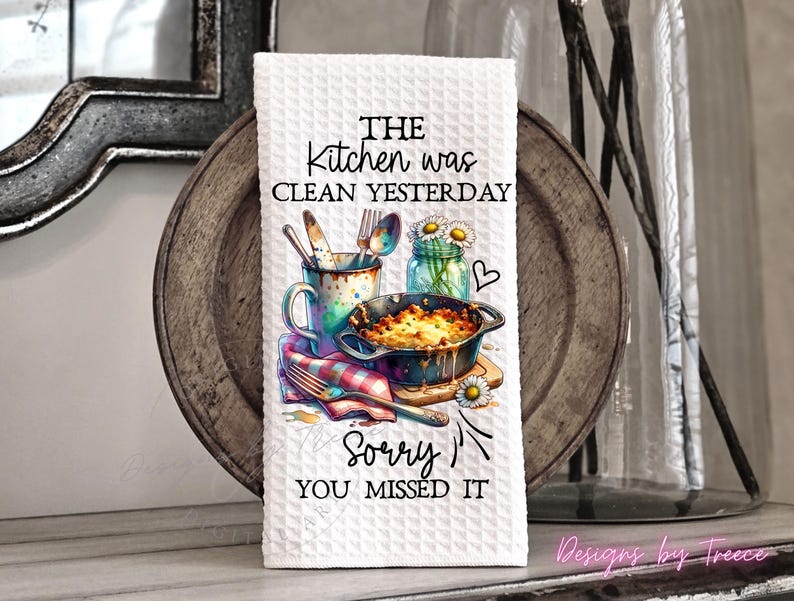 May include: White kitchen towel with a waffle weave texture. The towel features a colorful illustration of kitchen utensils, a casserole dish, and a jar of daisies. Text on the towel reads, "THE kitchen was clean yesterday, sorry you missed it."
