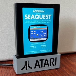 May include: A black Atari Seaquest video game cartridge with a blue label featuring game graphics. The cartridge is in a gray Atari-branded stand. The label reads "ACTIVISION PRESENTS SEAQUEST".