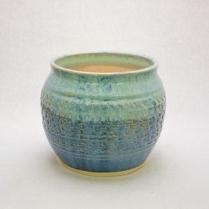 May include: A handcrafted ceramic planter with a textured, basket-weave design. The planter features a gradient of blue and teal hues, with a light beige interior. The round pot is suitable for indoor or outdoor use.