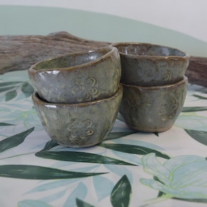 May include: Set of four small, hand-thrown ceramic bowls with a light green glaze and a subtle floral pattern. The bowls are stacked on top of each other.