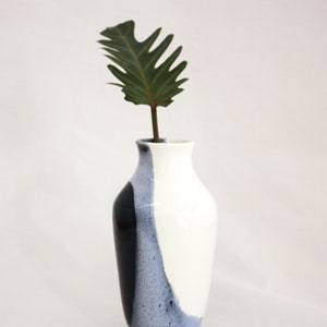 May include: A white ceramic vase with a black and blue glaze design. The vase has a rounded body and a narrow neck. A single green leaf is in the vase.