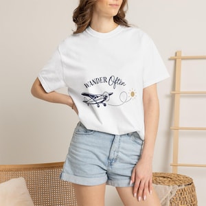 Puede incluir: Camiseta blanca con la frase "Wander Often" y un dibujo de avión azul marino. Una línea de puntos conduce a un sol amarillo. La camiseta se combina con pantalones cortos vaqueros.