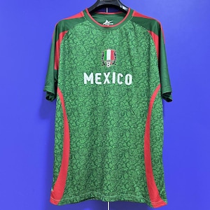 Può includere: Maglia da calcio verde con dettagli rossi e la scritta "MEXICO" in bianco. La maglia presenta uno stemma con i colori della bandiera messicana e un pallone da calcio. La maglia è appesa a una gruccia nera su sfondo blu.