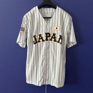 Può includere: Maglia da baseball bianca con strisce verticali nere, con la scritta "JAPAN" in lettere nere sul petto. Una toppa con la bandiera giapponese è sul petto destro e una toppa con testo giapponese sulla manica sinistra.
