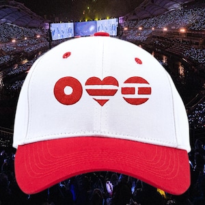 Può includere: Un cappellino da baseball bianco con visiera rossa e simboli rossi ricamati. I simboli includono un cerchio, un cuore e un rettangolo. Il cappellino è ambientato sullo sfondo di una grande folla a un concerto.