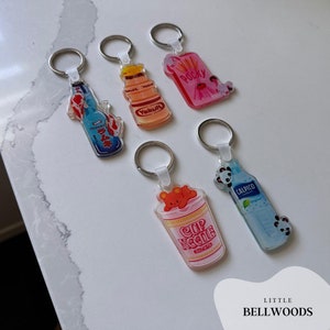 Pode incluir: Cinco chaveiros coloridos com populares lanches e bebidas japonesas. Os chaveiros são feitos de acrílico transparente e apresentam imagens de Yakult, Poccky, Calpico, Ramune e Cup Noodles.