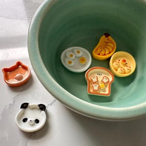 May include: A set of five small ceramic dishes featuring various breakfast foods. The dishes are shaped like a slice of toast, a banana, a fried egg, a panda face, and a fox face. The dishes are all white with colorful details.
