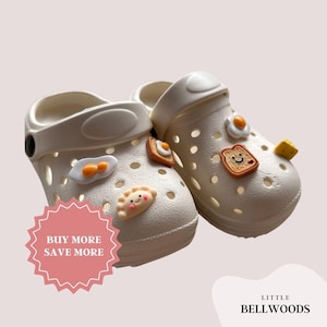 May include: A pair of off-white clogs decorated with food-themed charms. The charms include fried eggs, toast, a dumpling, and a cheese cube. The clogs have a strap and are perforated with small holes. The image also contains the text "BUY MORE SAVE MORE".
