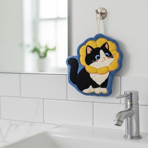May include: A cat-shaped sponge with a blue backing and a yellow, flower-like border. The cat is black and white with blue eyes. It hangs from a hook above a white sink.