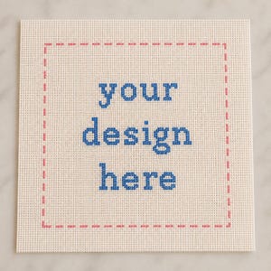 May include: A square piece of off-white Aida fabric featuring a cross-stitch border in pink. The center of the fabric displays the text your design here stitched in blue thread. The fabric has a visible grid pattern throughout the entire surface.