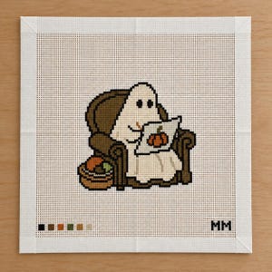 May include: A needlepoint canvas featuring a white ghost sitting in a dark brown armchair, reading a book with an orange pumpkin illustration on the cover. A small basket with orange and green items sits beside the chair. The letters MM are in the corner.