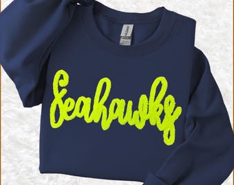 May include: A navy blue long-sleeve crewneck sweatshirt featuring the word Seahawks written in a bright neon yellow, textured, fuzzy chenille-style script font across the chest. The garment has a Gildan brand label at the inner neckline.