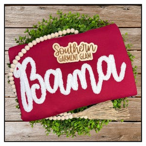 May include: A maroon sweatshirt featuring the word Bama in large white chenille letters. A small wooden tag above the text reads Southern GARMENT GLAM. The garment is surrounded by a circular wreath of green leaves and wooden beads.