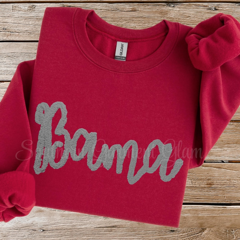 May include: A red long-sleeve crewneck sweatshirt featuring the word Bama in large, textured gray script lettering across the chest. The garment has a ribbed collar and cuffs, with a Gildan brand label visible inside the neckline.