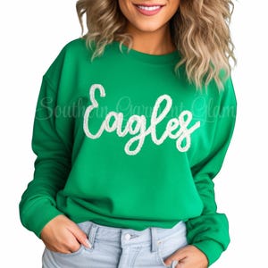 May include: A bright green crewneck sweatshirt featuring the word Eagles written in white, textured, fuzzy chenille-style script lettering across the chest. The garment has long sleeves with ribbed cuffs and a ribbed neckline.