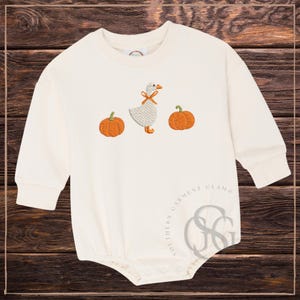 May include: A cream-coloured long-sleeve romper featuring an embroidered design of a white goose with an orange bow around its neck, positioned between two orange pumpkins. The garment has a round neckline and snap closures at the bottom.