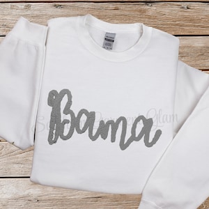 May include: A white long-sleeve crewneck sweatshirt featuring the word Bama written in a textured gray script font across the chest. The garment has a standard ribbed neckline and a Gildan brand label visible at the inner collar.