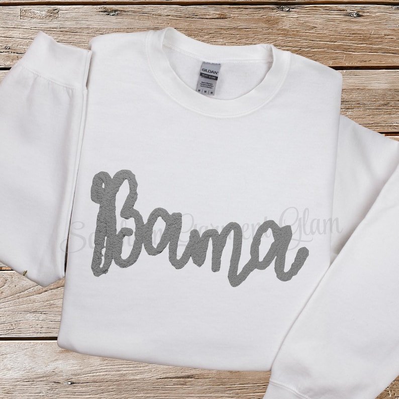 May include: A white long-sleeve crewneck sweatshirt featuring the word Bama written in a textured gray script font across the chest. The garment has a standard ribbed neckline and a Gildan brand label visible at the inner collar.