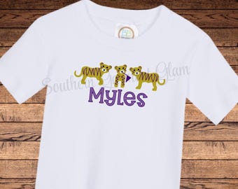 May include: A white short-sleeved t-shirt featuring an embroidered graphic of three gold tigers with brown stripes. The center tiger holds a small purple flag. Below the tigers, the name Myles is embroidered in purple block letters.