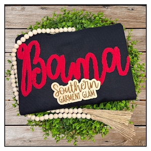 May include: A black t-shirt featuring the word Bama in large, red, textured chenille-style lettering. A small wooden tag is attached to the front with the text Southern Garment Glam engraved in a script and block font combination.