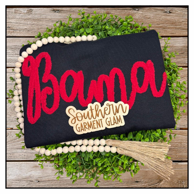 May include: A black t-shirt featuring the word Bama in large, red, textured chenille-style lettering. A small wooden tag is attached to the front with the text Southern Garment Glam engraved in a script and block font combination.