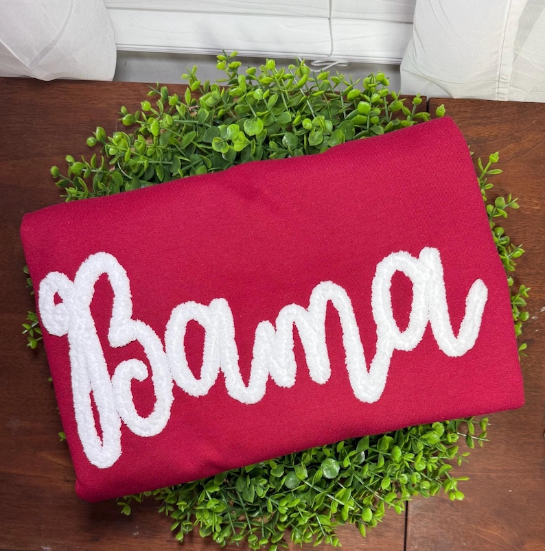 May include: A deep red sweatshirt featuring the word Bama written in large, white, textured chenille-style script lettering across the center. The garment is folded and placed on top of a circular bed of small, vibrant green artificial leaves.