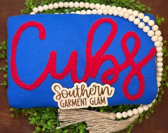 May include: A bright blue sweatshirt featuring the word Cubs in large, textured red chenille lettering. A wooden sign with the text Southern Garment Glam is placed over the fabric, and a string of light-colored wooden beads surrounds the item.