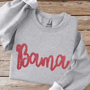 May include: A light gray crewneck sweatshirt featuring the word Bama written in a bold, textured red script font across the center. The garment has long sleeves, a ribbed neckline, and a Gildan brand label visible inside the collar.