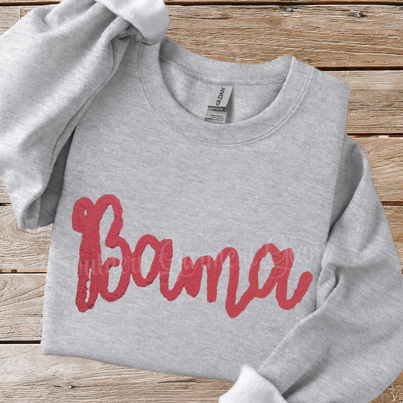 May include: A light gray crewneck sweatshirt featuring the word Bama written in a bold, textured red script font across the center. The garment has long sleeves, a ribbed neckline, and a Gildan brand label visible inside the collar.
