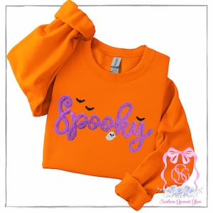 May include: A bright orange crewneck sweatshirt featuring the word Spooky embroidered in a textured purple script font. The design includes three small black bats and a tiny white ghost icon positioned below the lettering on the front chest.