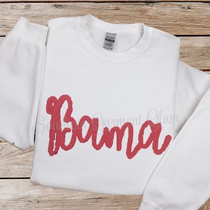 May include: A white long-sleeve crewneck sweatshirt featuring the word Bama written in a textured, coral-pink script font across the chest. The garment has a standard ribbed collar and a Gildan brand label visible at the inner neckline.