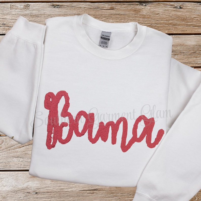 May include: A white long-sleeve crewneck sweatshirt featuring the word Bama written in a textured, coral-pink script font across the chest. The garment has a standard ribbed collar and a Gildan brand label visible at the inner neckline.